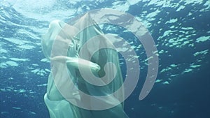 Young woman underwater model in white cloth on background of blue water.