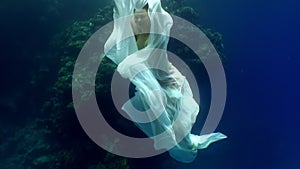 Young woman underwater model in white cloth on background of blue water.