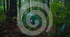 Young woman unzipping tent shining torch in green rainy forest outdoors. Wide shot Caucasian female tourist looking