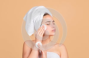 Young woman use micellar water and circle white pad, enjoys spa procedures, wrapped towel on head, isolated on beige photo