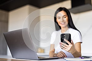 Young woman use phone sit at table in kitchen share text messages in social media, enjoy new vlog, watch internet content take