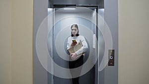 Young woman uses phone, holds cardboard box and getting out of the elevator, moving in to new appartment