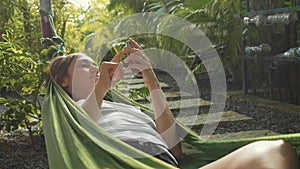 Young woman uses a smartphone while lying in a hammock at green garden.