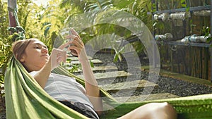 Young woman uses a smartphone while lying in a hammock at green garden