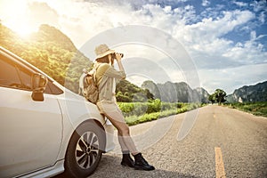 A young woman using black binoculars outside her car on the road of travel