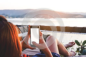 Young woman using black smartphone with blank desktop screen while lying side the sea with evening sunset background.