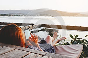 Young woman using black smartphone with blank desktop screen while lying side the sea with evening sunset background.