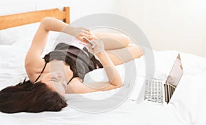 Young woman using cellphone and laptop in bed