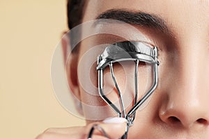 Young woman using eyelash curler on beige background, closeup