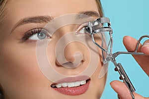 Young woman using eyelash curler on light blue background, closeup