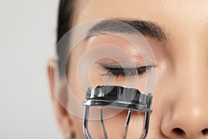 Young woman using eyelash curler on light grey background, closeup