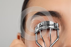 Young woman using eyelash curler on light grey background, closeup