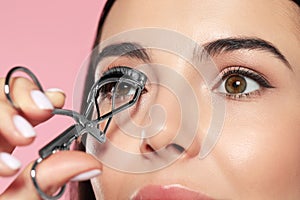 Young woman using eyelash curler on light pink background, closeup