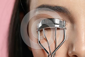 Young woman using eyelash curler on light pink background, closeup