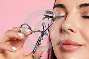 Young woman using eyelash curler on light pink background, closeup