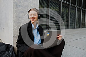 Young woman using headphones and laptop while sitting on the floor outdoors