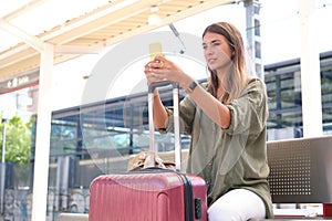 Young woman using her smartphone and resting upon her suitcase, waiting for the train at a railway station