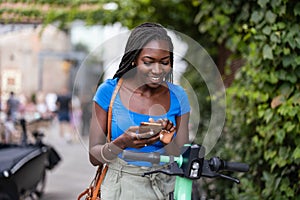 Young woman using her smartphone while standing with electric scooter in the city