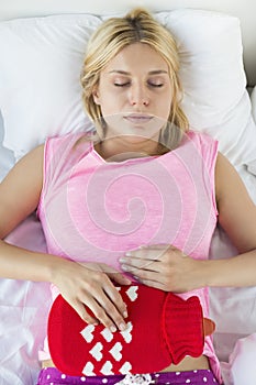 Young woman using hot water bottle on abdomen while lying on bed