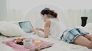 Young woman using laptop during breakfast lying on white bed at home. Brunette girl typing on pc computer.