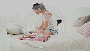 Young woman using laptop during breakfast sitting on white bed at home. Brunette girl typing message on pc, chatting.