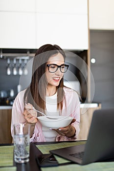 Young cheerful woman using on laptop computer and eating breakfast in the kitchen