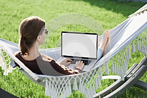 Young woman using laptop computer with white blank empty screen lying in hammock.