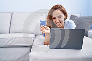 Young woman using laptop and credit card lying on sofa at home