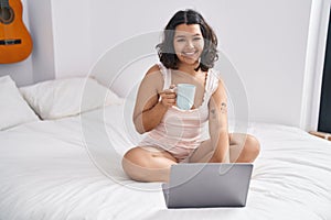 Young woman using laptop drinking coffee sitting on bed at bedroom