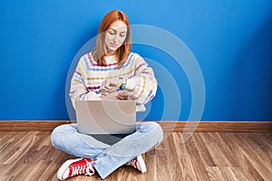 Young woman using laptop at home sitting on the floor checking the time on wrist watch, relaxed and confident