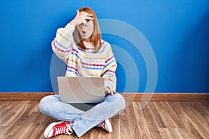 Young woman using laptop at home sitting on the floor peeking in shock covering face and eyes with hand, looking through fingers