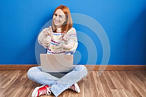 Young woman using laptop at home sitting on the floor pointing to you and the camera with fingers, smiling positive and cheerful