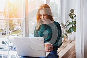 Young woman using laptop at home, typing, studying, watching webinars and drinking coffee