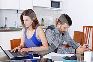 Young woman using laptop and man with phone at home kitchen