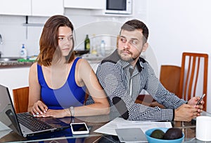 Young woman using laptop and man with phone at home kitchen