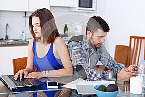 Young woman using laptop and man with phone at home kitchen