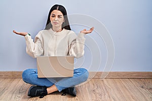 Young woman using laptop sitting on the floor at home clueless and confused expression with arms and hands raised