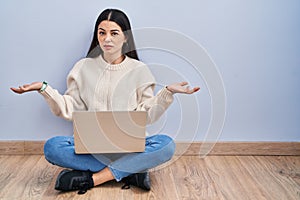 Young woman using laptop sitting on the floor at home clueless and confused with open arms, no idea concept