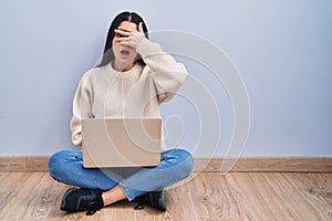 Young woman using laptop sitting on the floor at home peeking in shock covering face and eyes with hand, looking through fingers