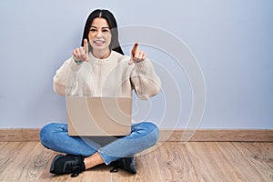 Young woman using laptop sitting on the floor at home pointing to you and the camera with fingers, smiling positive and cheerful