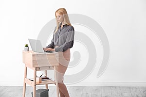 Young woman using laptop at stand up workplace against white wall.