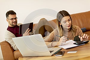 Young woman using laptop and young man using digital tablet while sitting on sofa at home