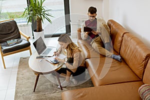 Young woman using laptop and young man using digital tablet while sitting on sofa at home