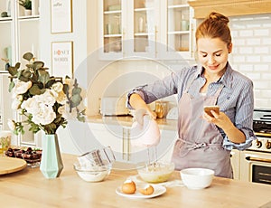 Young woman using mixer standing in kitchen and looking at recipe on mobile phone