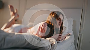 Young woman using mobile phone at home. Smiling happy female lying in bed typing