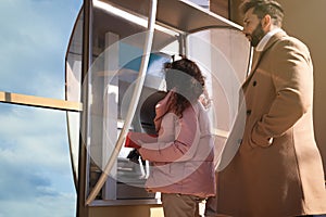 Young woman using modern cash machine outdoors. ATM queue