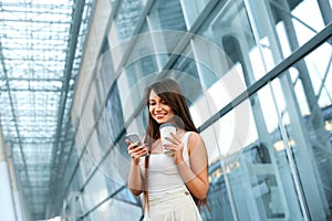 Young Woman Using Phone Outdoors. Portrait Of Beautiful Business Woman Holding Smartphone And Cup Of Coffee In Hands