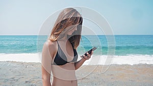 Young woman using smartphone on beach on summer travel vacation by sea. She is wearing bikini while relaxing during