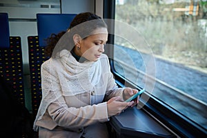 Young woman using smartphone while enjoying her business travel by train, sitting on seat by window overlooking downtown