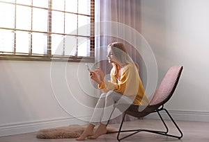 Young woman using smartphone near window with blinds